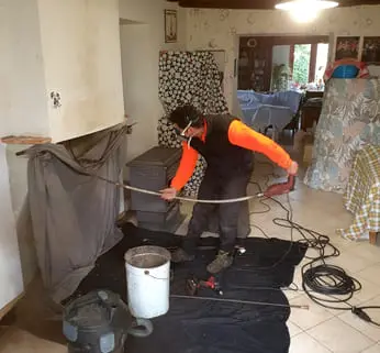 Ramoneur nettoyant le conduit d'une cheminée depuis le foyer, sol protégé par une bâche